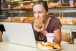 Young woman eating pastry stressed in front of computer